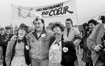 Coluche pose avec deux bénévoles devant l'entrée du Restaurant du Cœur à Gennevilliers pour l'inauguration d'un des trois Restaurants du Cœur de la région parisienne, le 21 décembre 1985 © Michel Gangne / AFP